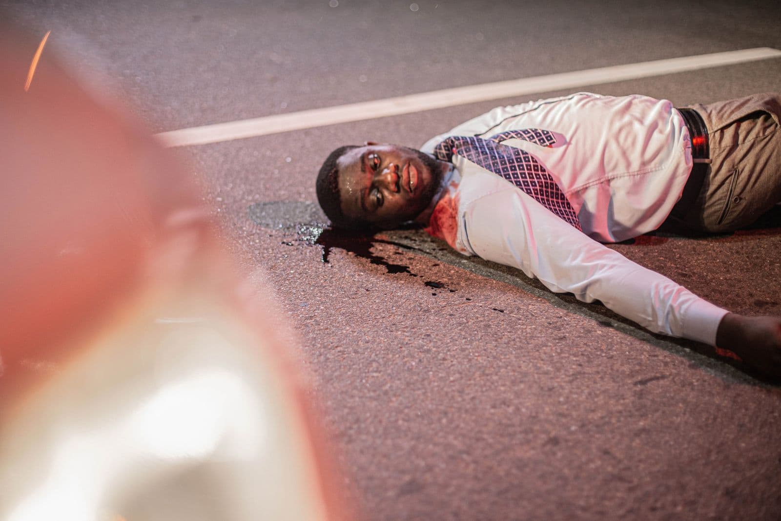 A man lying on a road beside a car, a staged crash scene